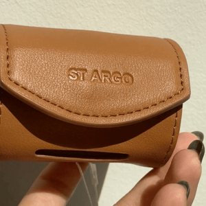 A close-up of a caramel vegan leather St. Argo poop bag holder being held by a hand with dark nail polish, against a light textured wall.