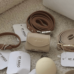 A styled flat-lay of St.argo accessories on a beige textured rug, including a caramel leather lead, and a cream poop bag holder with white brand tags.
