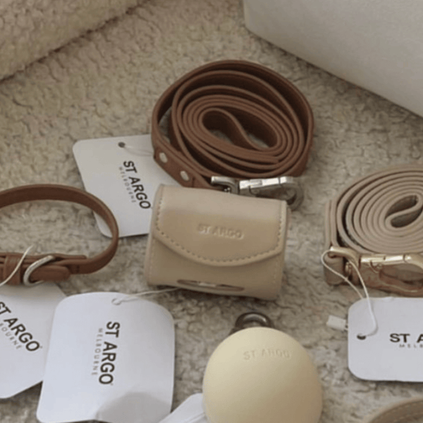 A styled flat-lay of St.argo accessories on a beige textured rug, including a caramel leather lead, and a cream poop bag holder with white brand tags.