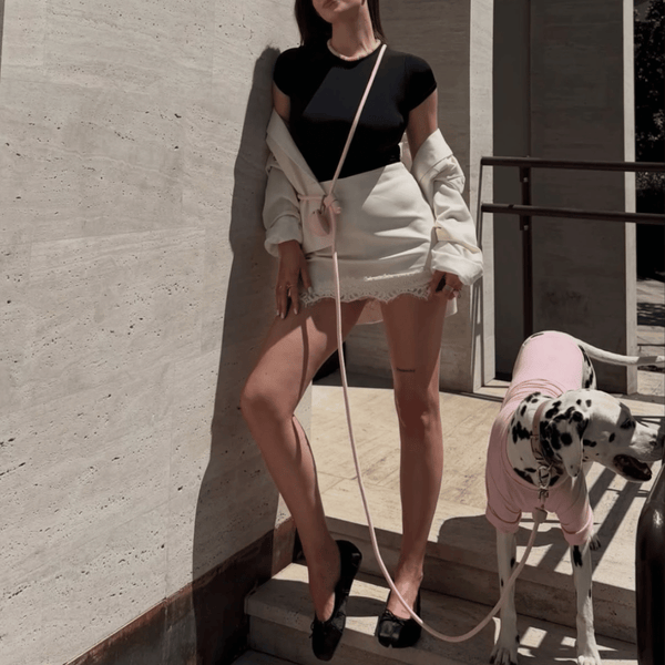 Woman in black top and white shorts standing next to a Dalmatian dog in a pink outfit against a concrete wall with the marshmallow hands-free lead across her body.