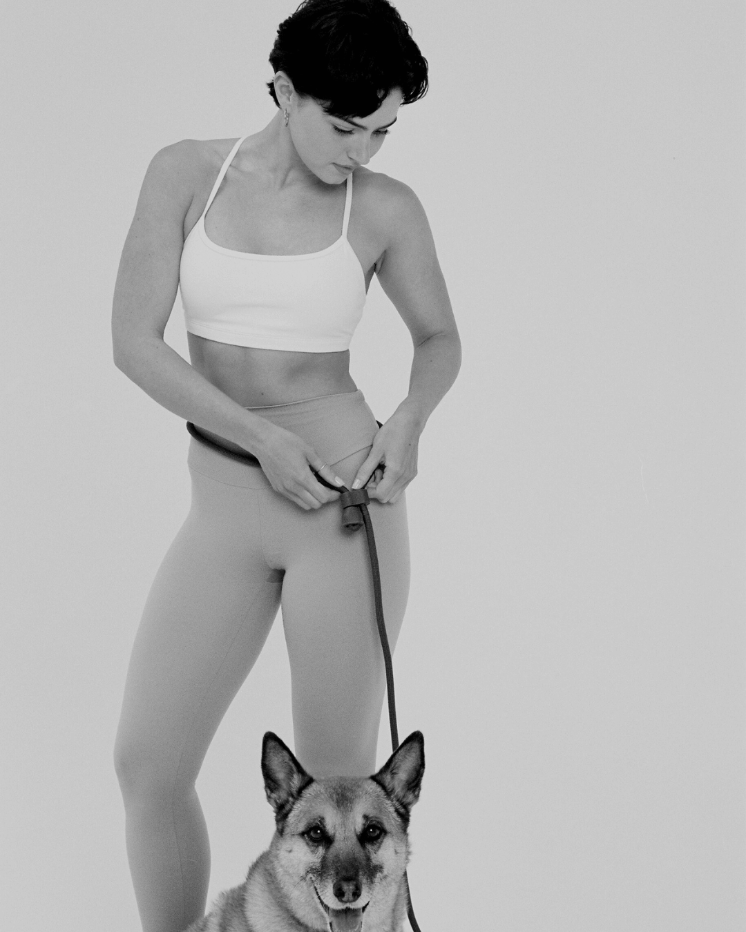 Woman in athletic wear holding a dog leash with a dog at her feet on a plain background